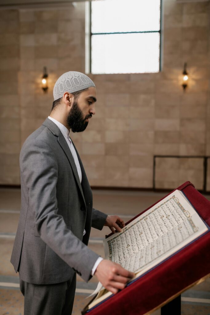 A Muslim man in a gray suit reads the Quran at a mosque, emphasizing Islamic culture and spirituality.